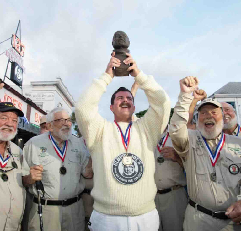 Surrounded by previous winners of the Hemingway Look-Alike Contest, 2026 champion Chris Dutton hoists his trophy, a bronze bust of Nobel Prize-winning author Ernest Hemingway, outside Sloppy Joe’s Bar.